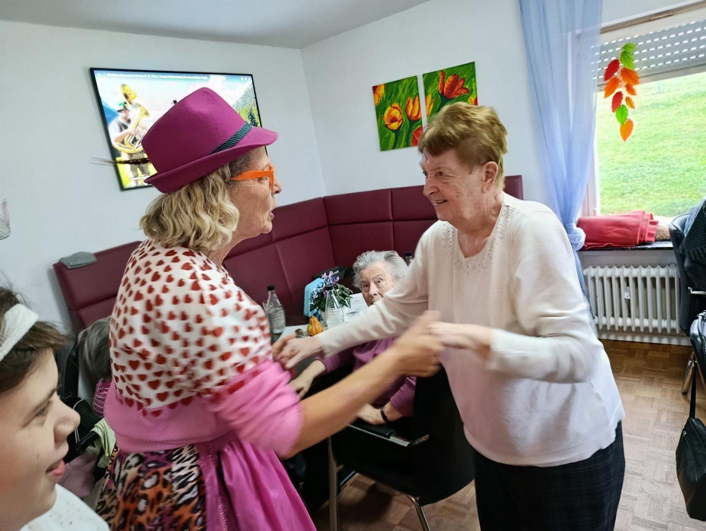 Drei Frauen sitzen in einem Wohnzimmer und unterhalten sich, wobei eine Dame einen auffälligen Hut trägt.