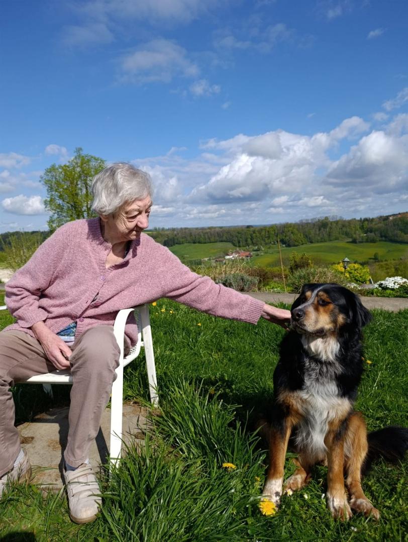 Eine ältere Frau in einem rosafarbenen Pullover sitzt draußen auf einem Stuhl neben einem großen, sitzenden Hund vor einer weiten grünen Landschaft.