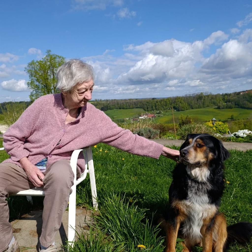 Eine ältere Frau in einem rosafarbenen Pullover sitzt draußen auf einem Stuhl neben einem großen, sitzenden Hund vor einer weiten grünen Landschaft.